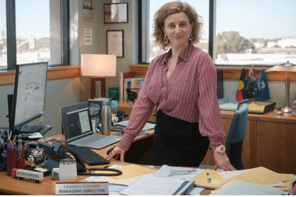 Felicity Ward como Hannah Howard sentada em uma mesa de um escritório em "The Office"