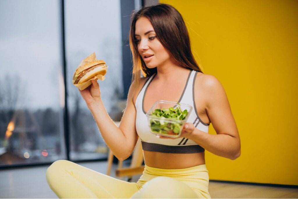 Mulher olhando para um hambúrguer e segurando pote de salada