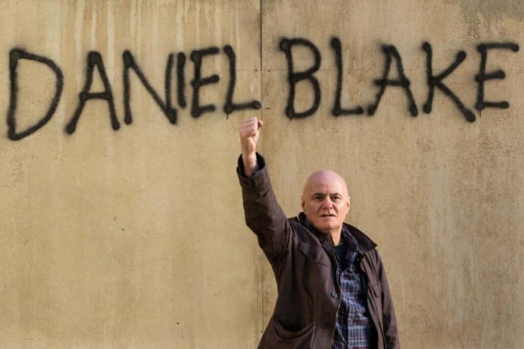 Homem em frente a uma parede com grafite com um dos braços levantados para o alto em cena do filme  "Eu, Daniel Blake" 