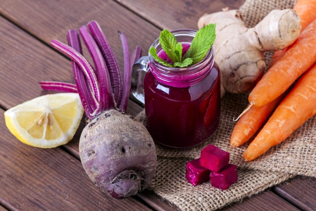 Suco de beterraba, cenoura, gengibre e limão, servido em jarro, cercado pelos ingredientes frescos sobre uma superfície de madeira