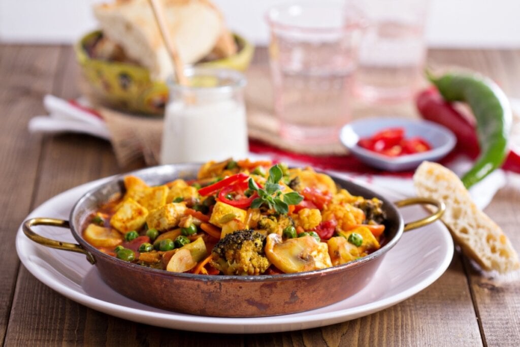 Prato de curry vegano com tofu, legumes frescos e pimentas, servido em uma panela de cobre