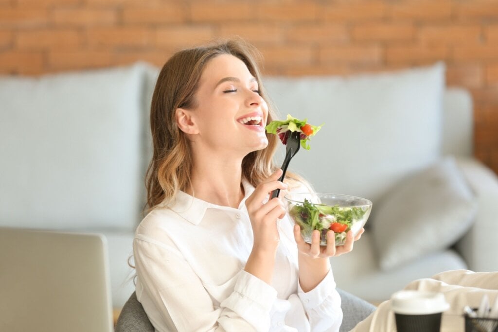 Mulher segurando pote de salada e levando garfo com salada á boca