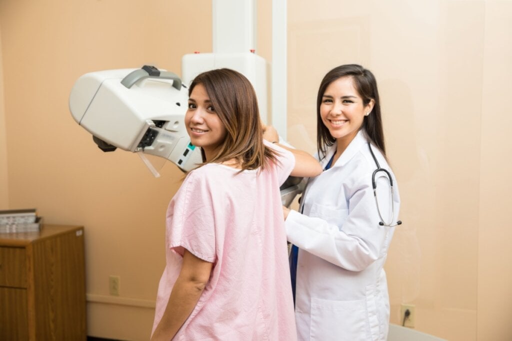 Mulher em sala de exame realizando mamografia acompanhada de uma médica.