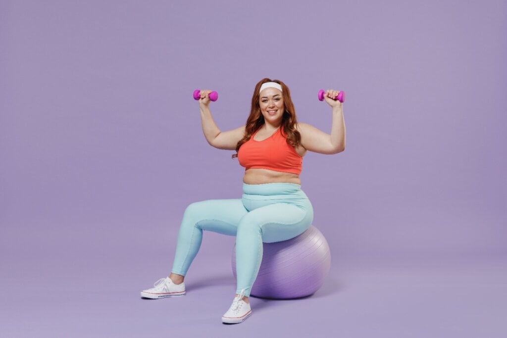 Mulher acima do peso segurando halteres e sentada em bola de pilates