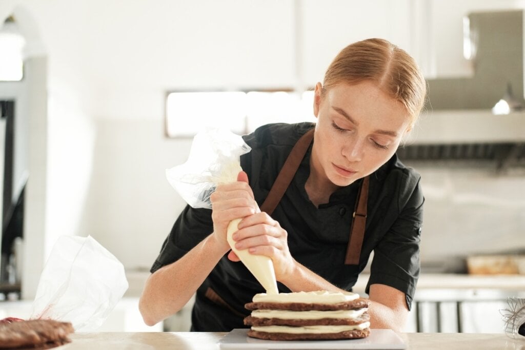 Mulher em uma cozinha confeitando um bolo com um saco de confeiteiro 