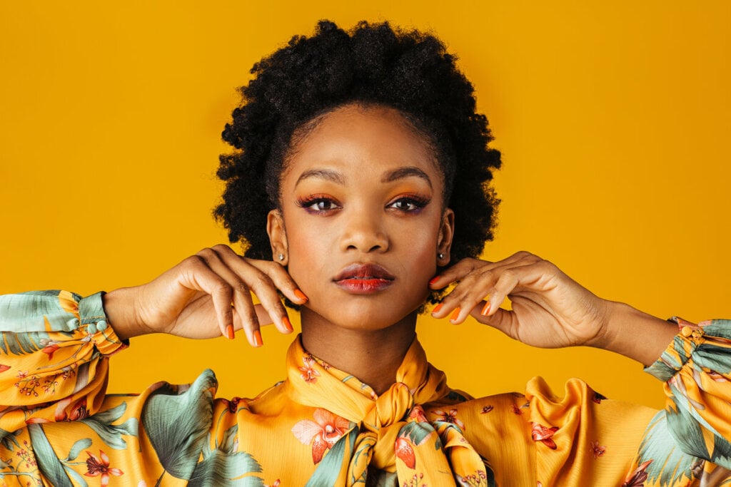 Mulher maquiada vestindo blusa amarelo floral em frente a um fundo amarelo