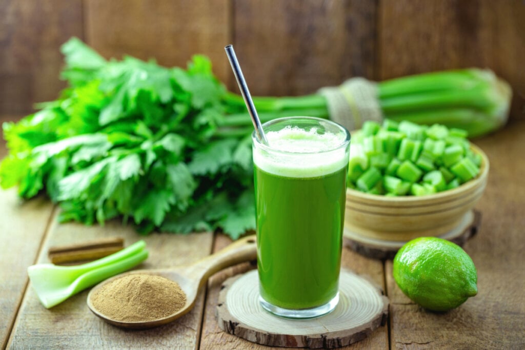 Copo de suco verde com canudo de metal. Ao fundo, aipo, canela e limão