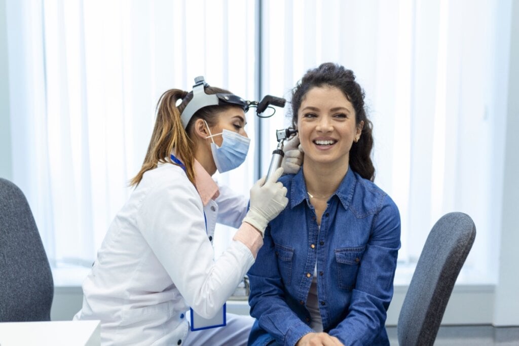 Paciente sorridente recebendo atendimento de uma médica otorrinolaringologista, que examina sua orelha usando um otoscópio