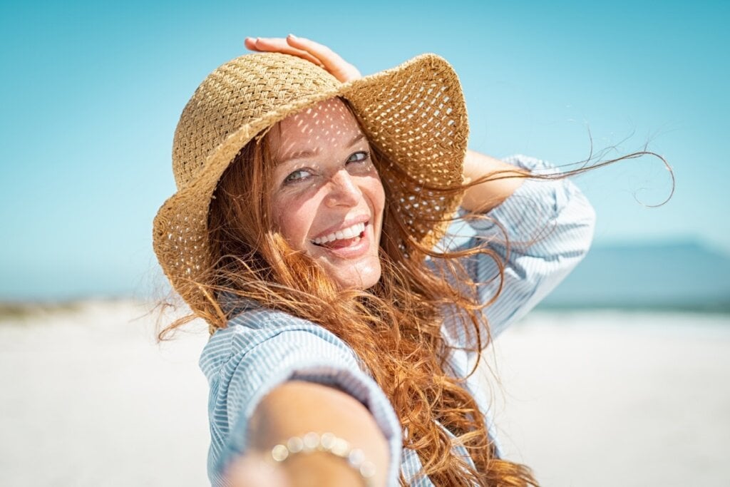 Mulher ruiva, sorrindo e usando um chapéu em uma praia 