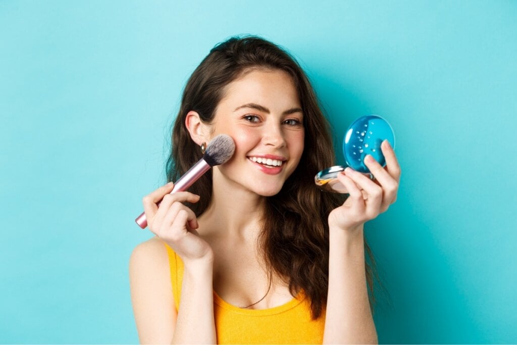 Mulher sorrindo, usando uma blusa amarela e posando para foto em um fundo azul-claro maquiando o rosto