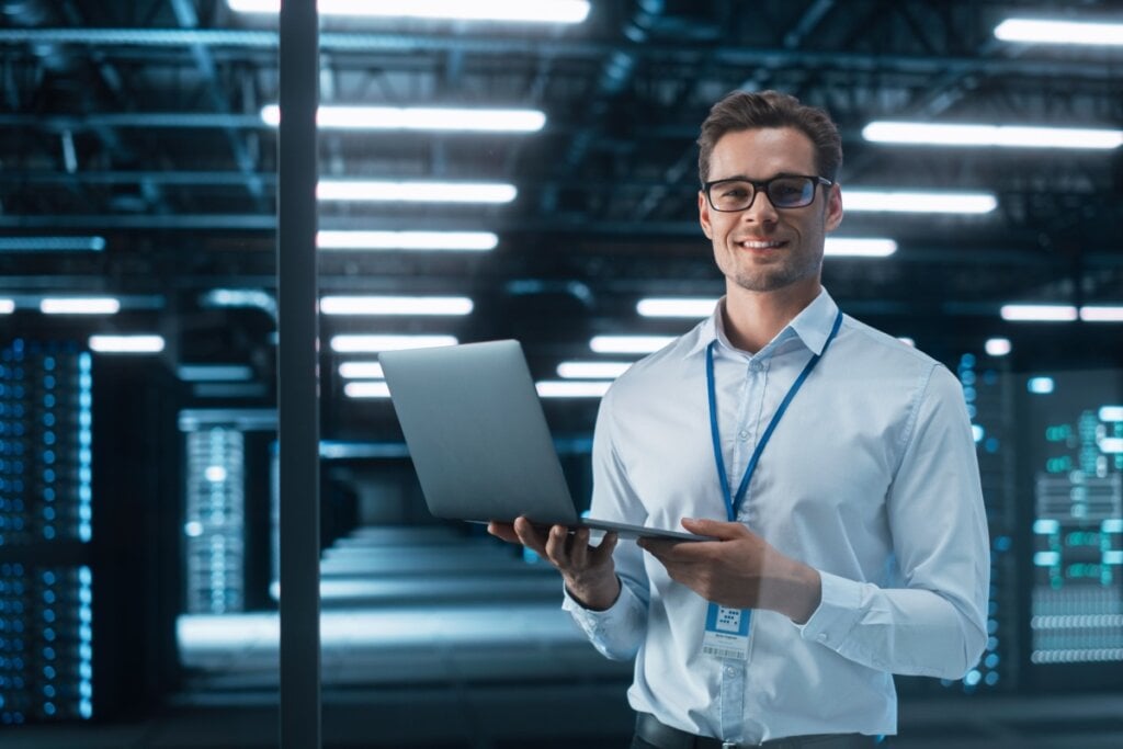 Homem sorridente segurando um notebook em um ambiente de data center