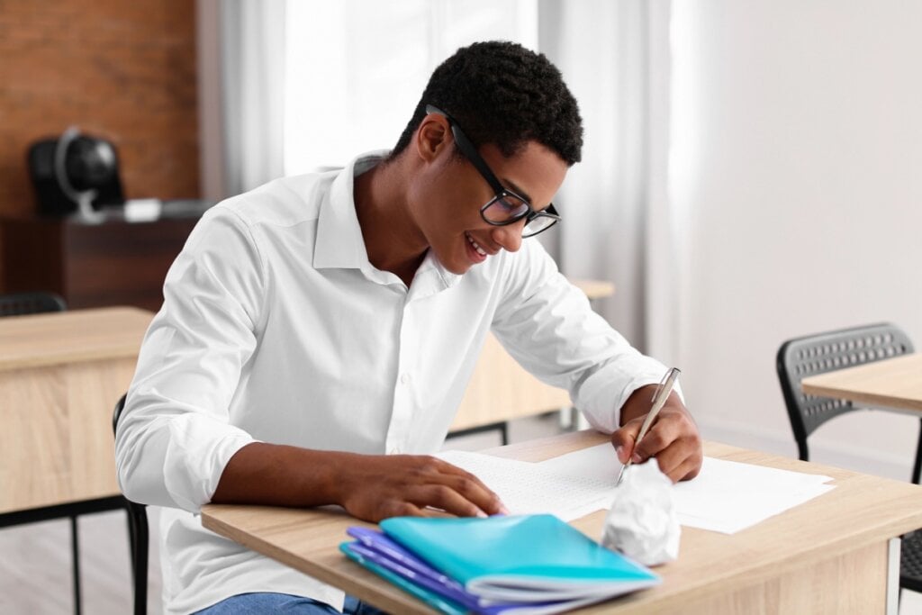 Estudante estudando; em cima da mesa, há apostilas e papéis