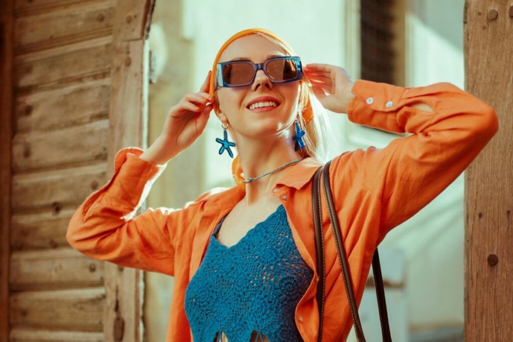 Mulher usando um lenço laranja na cabeça, blus a azul, camisa laranja, óculos de sol e brincos em formato de estrela sorrindo 