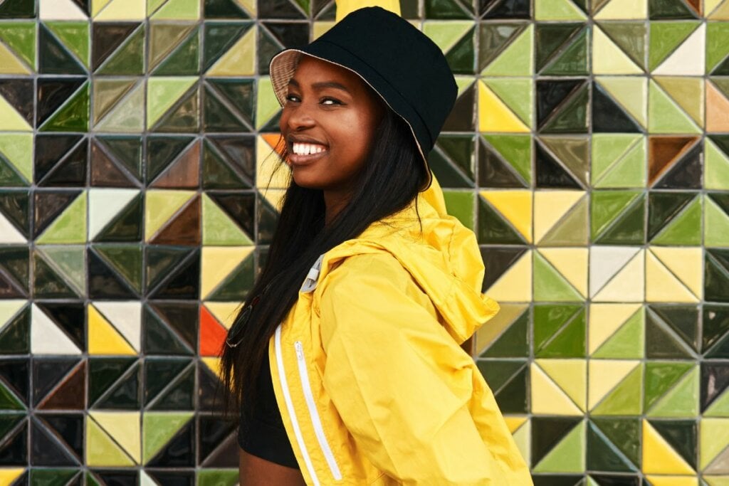 Mulher sorrindo e usando um chapéu bucket preto em frente a uma parede com azulejos coloridos 