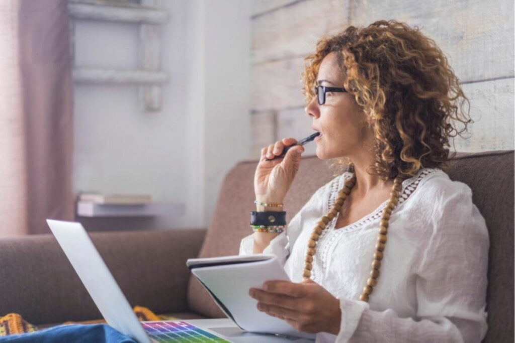 Mulher sentada com um notebook no colo e segurando um bloco de notas pensando 