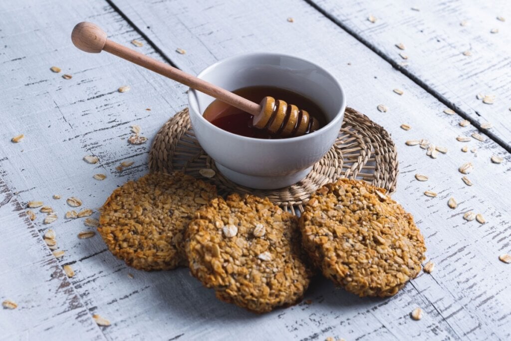 Biscoitos de aveia com chia, servidos em cima de mesa de madeira ao lado de tigela de porcelana com mel 