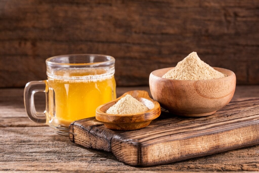 Caneca de vidro com chá de maca peruana, e ao lado duas cumbucas de madeira com maca peruana em pó em cima de mesa de madeira