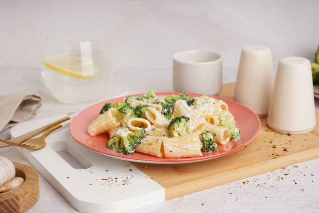 prato laranja com macarrão rigatoni com molho branco e brócolis. há potinhos brancos de tempero ao lado