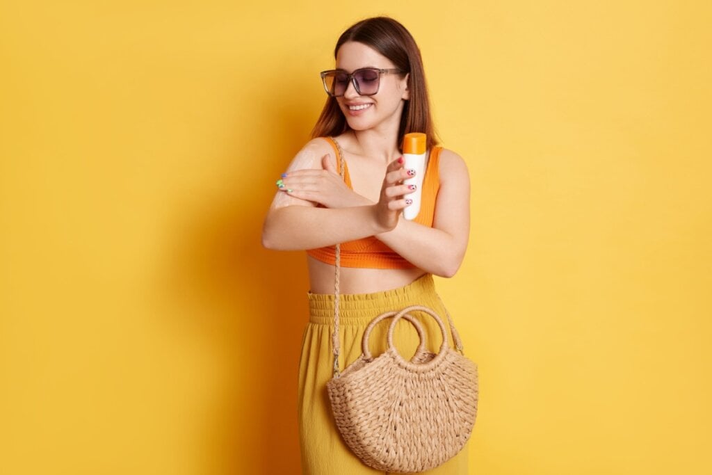 mulher de óculos de sol, camiseta laranja e calça amarela aplicando protetor solar