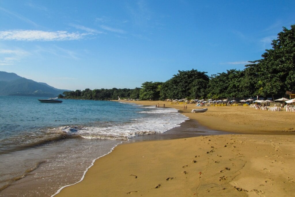 Praia do Curral, em Ilhabela, com ondas, sombreiros e vegetação verde abundante 