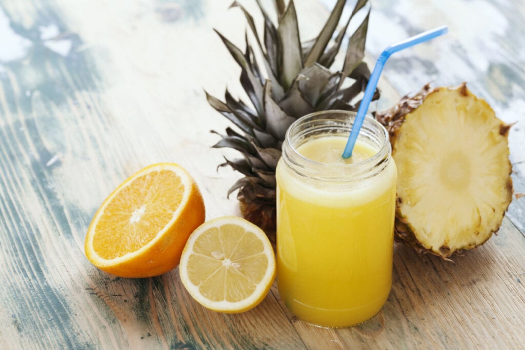 Copo de vidro com suco amarelo e canudo azul. Em volta, abacaxi, laranja e limão-siciliano