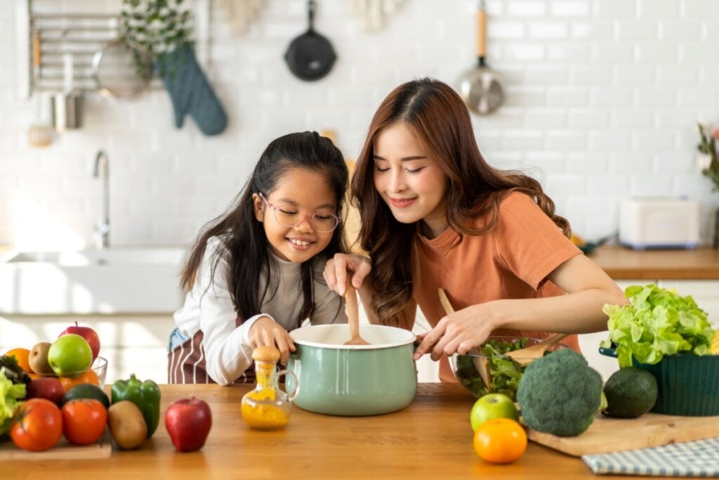 Mulher e uma menina em uma cozinha, mexendo em uma panela verde em cima de uma mesa de madeira, com frutas e vegetais ao redor