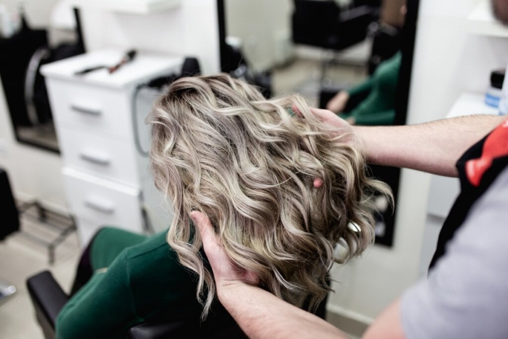 Pessoa com as mãos no cabelo loiro e liso de uma mulher sentada de costas 