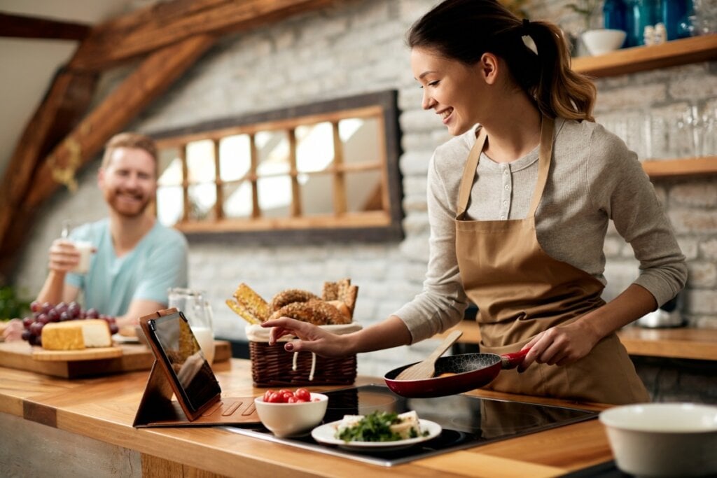 Mulher em uma cozinha cozinhando ao lado de um homem sorrindo ao seu lado tomando suco