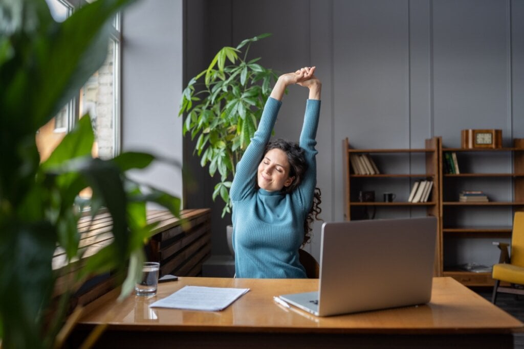 Mulher sentada em frente a uma mesa com notebook alongando os braços 