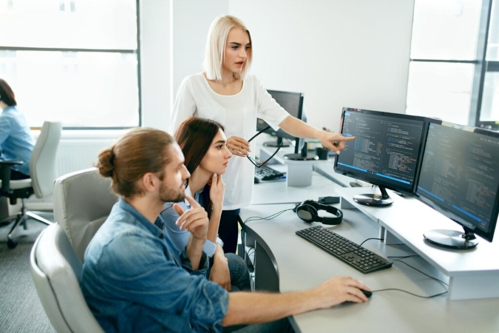 Homem e mulher sentados em frente a um computador e uma mulher em pé do lado apontando para a tela do aparelho