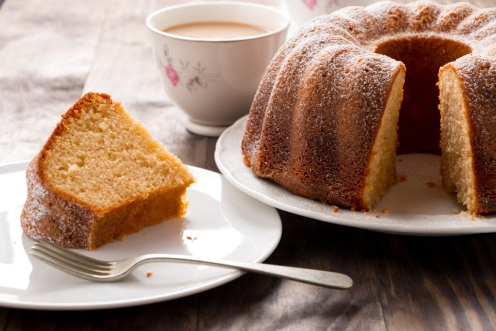 Bolo de iogurte com fatia servida em prato de cerâmica branco e com xícara de café atrás