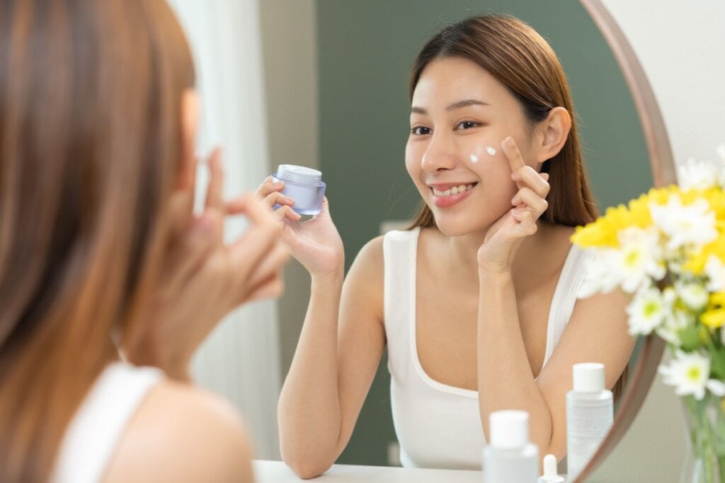 mulher se olhando no espelho, segurando potinho de creme e aplicando produto no rosto