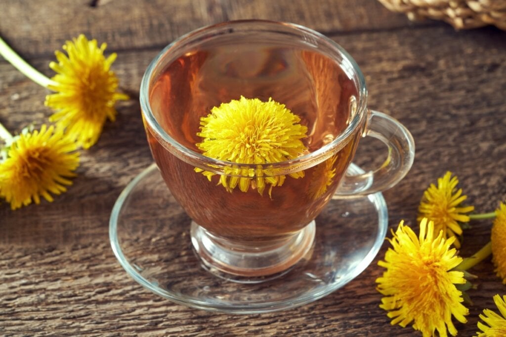 Chá de dente-de-leão servido em xícara de vidro em cima de mesa de madeira, com flores de dente-de-leão ao redor