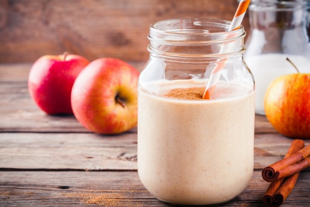 9 vitaminas caseiras para desintoxicar o organismo 6 Copo de vidro com vitamina de maçã decorada com canela em pó em cima de uma mesa de madeira com maçãs