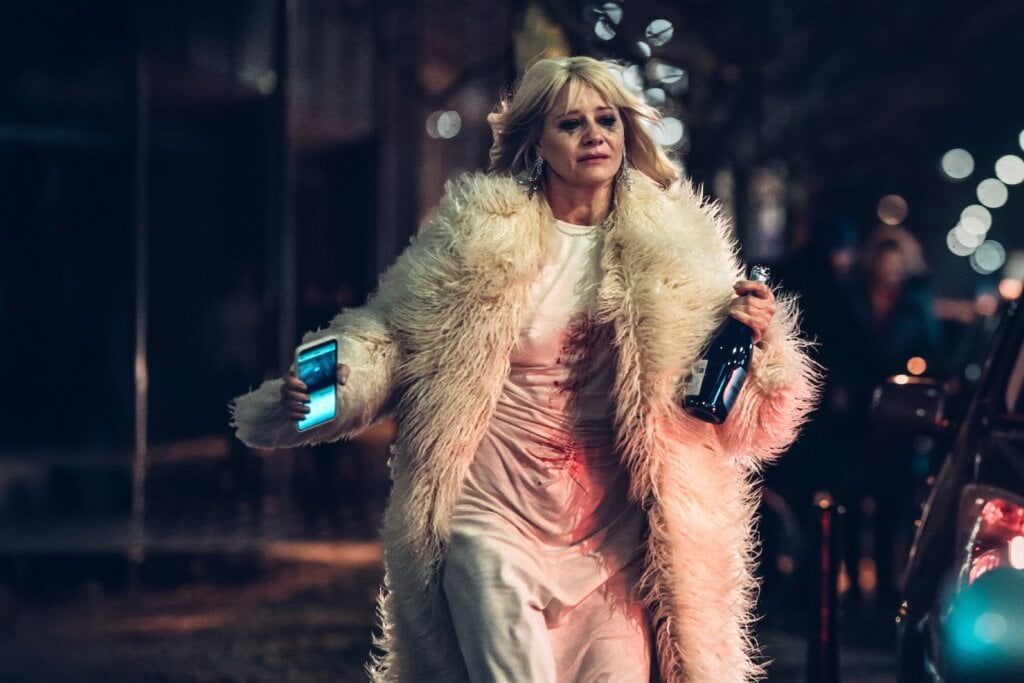 5 lançamentos imperdíveis da Netflix em junho de 2025 23 Mulher usando um casaco peludo e vestido, andando na rua, segurando o celular e uma garrafa de champanhe