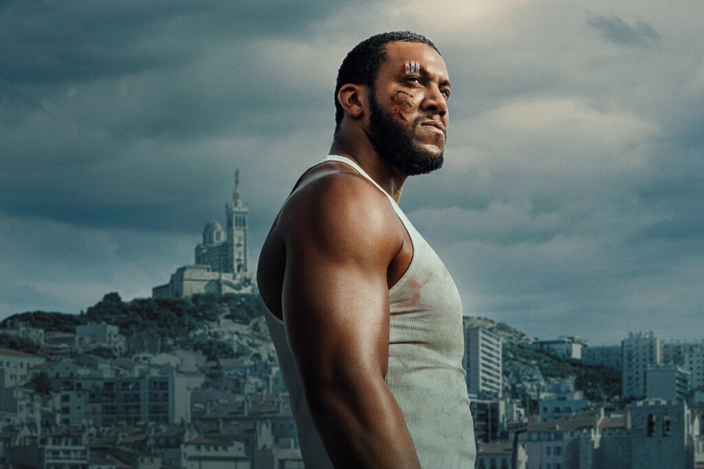 5 lançamentos imperdíveis da Netflix em junho de 2025 21 Homem em pé, com o rosto com um curativo, usando regata e com a imagem de uma cidade ao fundo