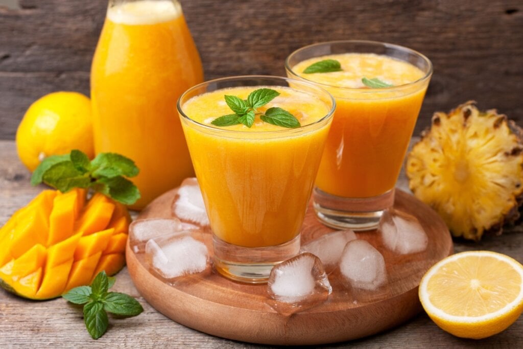 7 sucos cítricos para ajudar a controlar a pressão alta 6 Dois copos de vidro com suco de manga, abacaxi e hortelã em descanso de madeira ao lado de manga, abacaxi, laranja cortados e folhas de hortelã