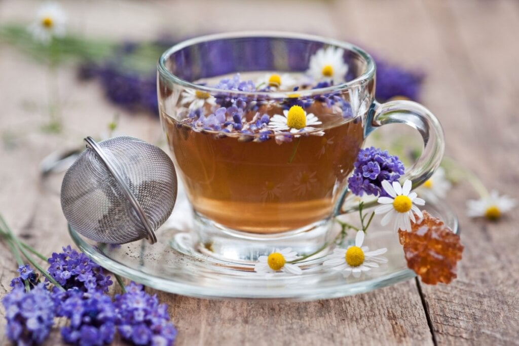 7 chás de camomila e seus benefícios para a saúde 6 xícara de vidro com chá com folhas de camomila e lavanda. ao lado da xícara, mais folhas de camomila e lavanda