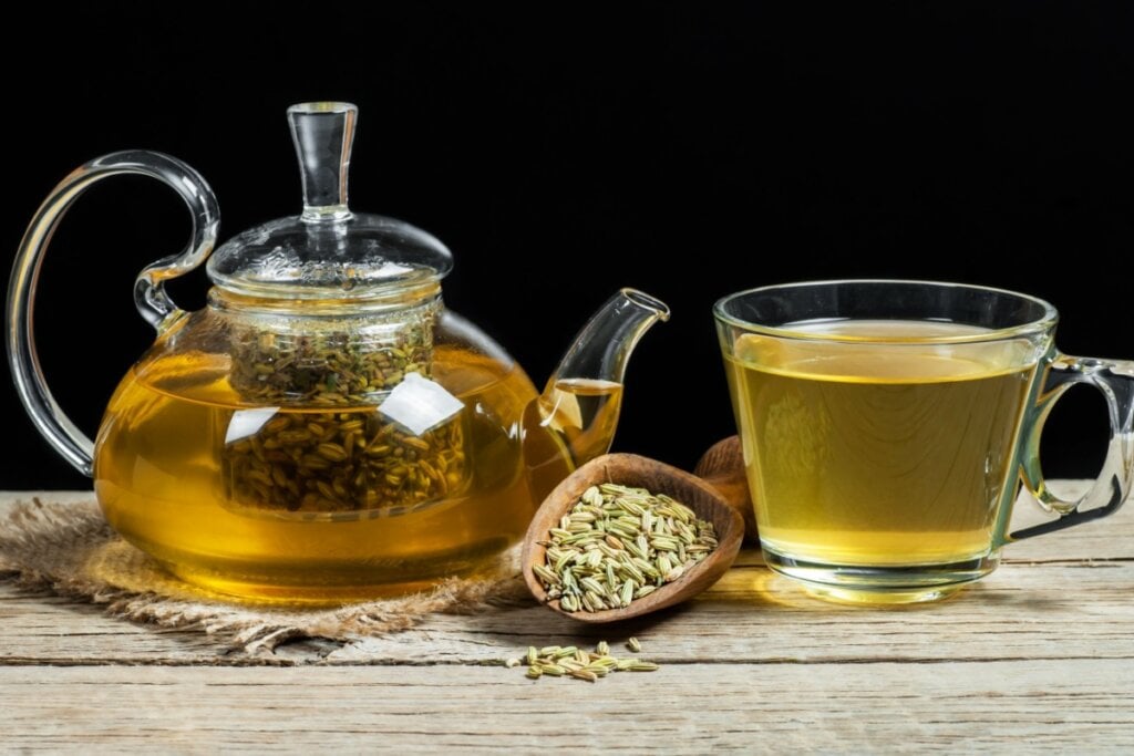 13 chás para melhorar a saúde e ajudar a emagrecer 6 Chá de erva-doce em bule de vidro e xícara de vidro, com uma colher de madeira com sementes de erva-doce, ambos em cima de uma mesa de madeira