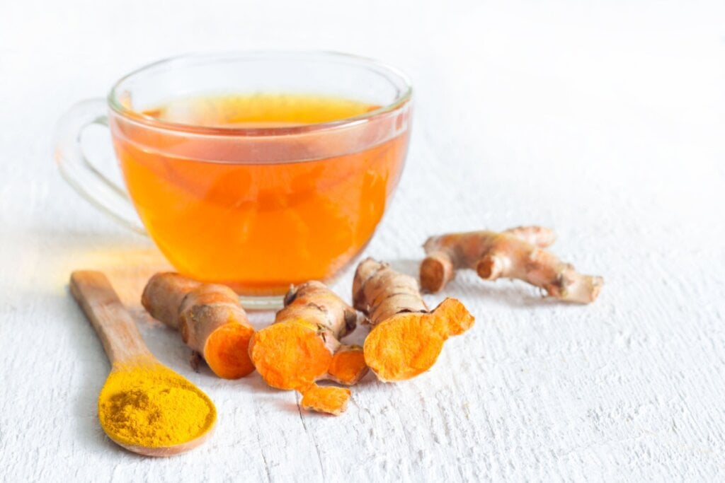 7 chás para melhorar a saúde do fígado 6 Chá de cúrcuma servido em xícara de vidro com pedaços de cúrcuma ao lado e uma colher de pau com cúrcuma em pó