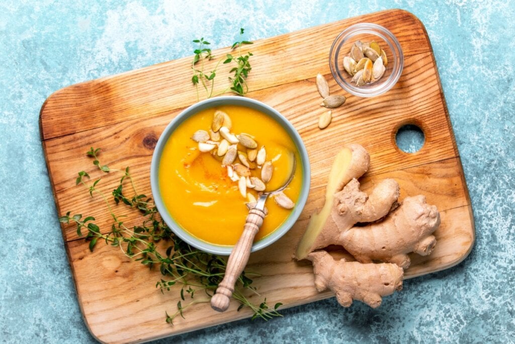 7 caldos e cremes veganos para os dias frios 6 Creme de abóbora com gengibre servido em tigela de cerâmica azul, com colher de metal e madeira, servido em cima de tábua de madeira em cima de uma mesa de pedra azul, ao lado estão pedaços de gengibre e sementes de abóbora em uma tigela