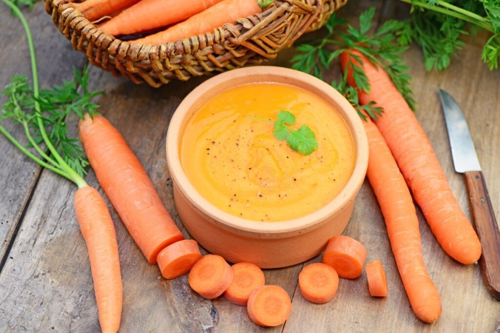 8 receitas de sopas e cremes termogênicos para o outono 6 Creme de cenoura com cúrcuma servido em tigela de cerâmica em cima de mesa de madeira e com cenouras ao lado
