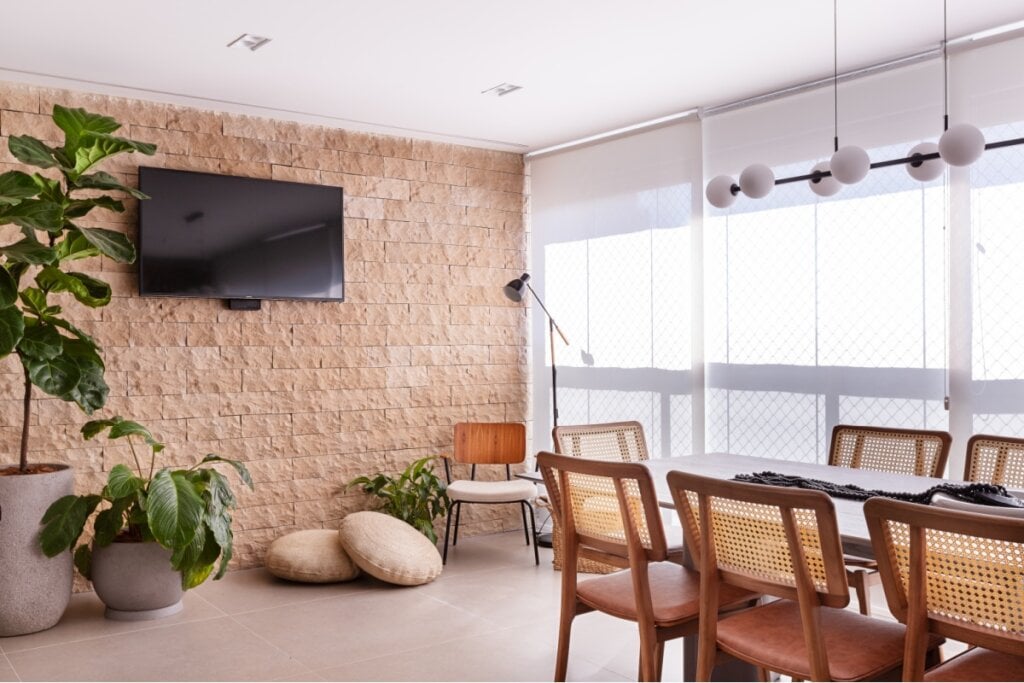 3 materiais que elevam a atmosfera dos ambientes 12 Sala de jantar com uma parede com pedra natural marrom e, a frente dela, uma mesa de madeira com cadeiras e uma janela com cortinas ao lado