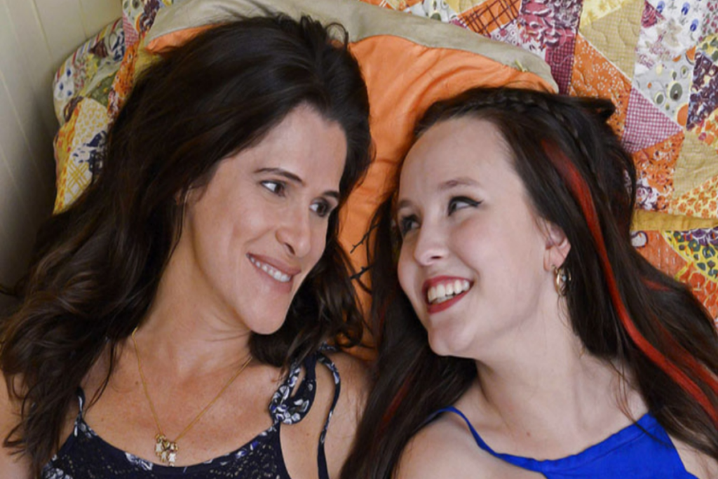 6 filmes de comédia sobre mães brasileiras 28 Mulher com cabelo preto liso solto e usando regata de alcinha deitada ao lado de uma adolescente com o cabelo preto e com mecha colorida usando regata azul