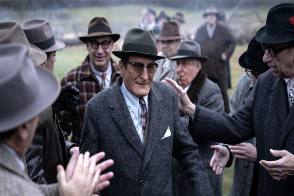 5 filmes e séries que estreiam na Max em junho de 2025 23 Homem no centro usando terno cinza, camisa branca, óculos e chapéu cerdo de outros homens também com roupas sociais