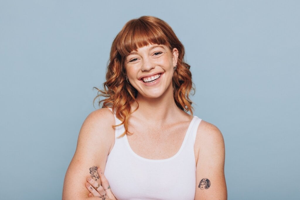 5 cores para transformar o cabelo para o inverno 24 Jovem com o cabelo curto e com franja ruivos, usando regata branca em fundo azul, está sorrindo e com os braços cruzados