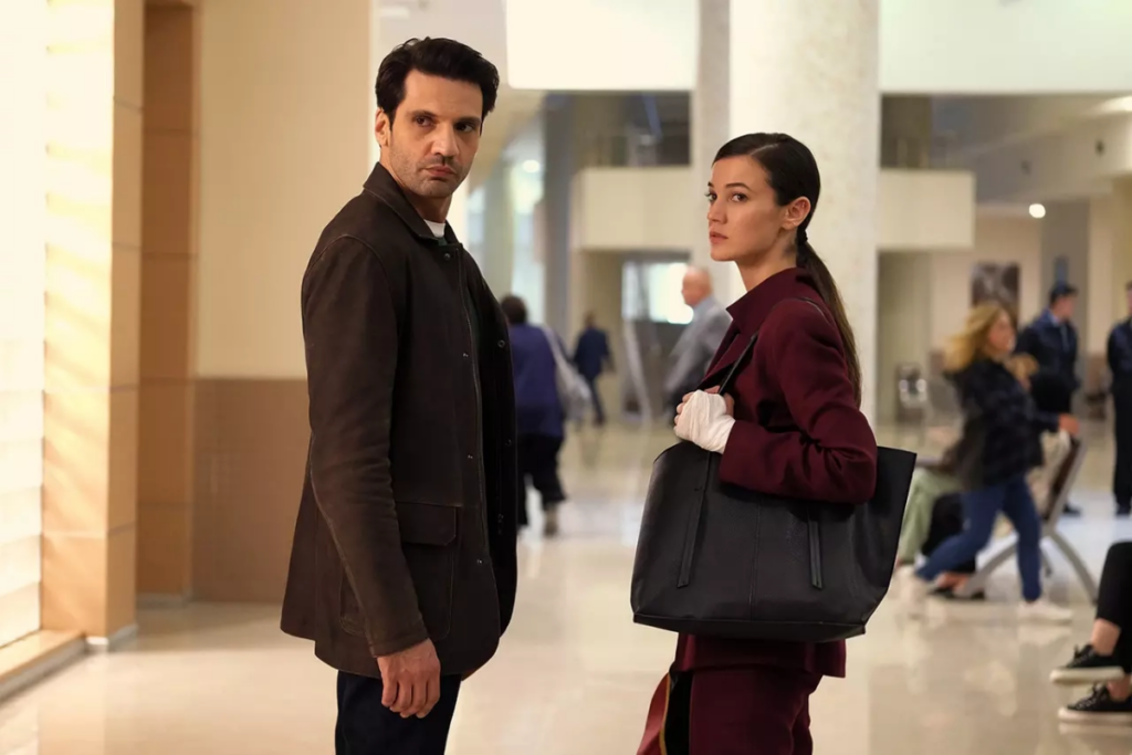 5 filmes e séries que estreiam na Max em junho de 2025 21 Homem com cabelo curto, usando blazer marrom e calça e blusa preta parado ao lado de uma mulher com o cabelo amarrado em rabo de cavalo e usando uma roupa social vinho com um curativo na mão e com uma bolsa preta