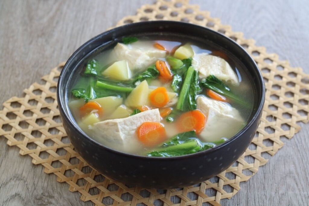 8 sopas deliciosas para ajudar a emagrecer 6 Sopa de tofu com cenoura, batata e espinafre servido em cumbuca de cerâmica preta em cima de descanso de panela em mesa de madeira