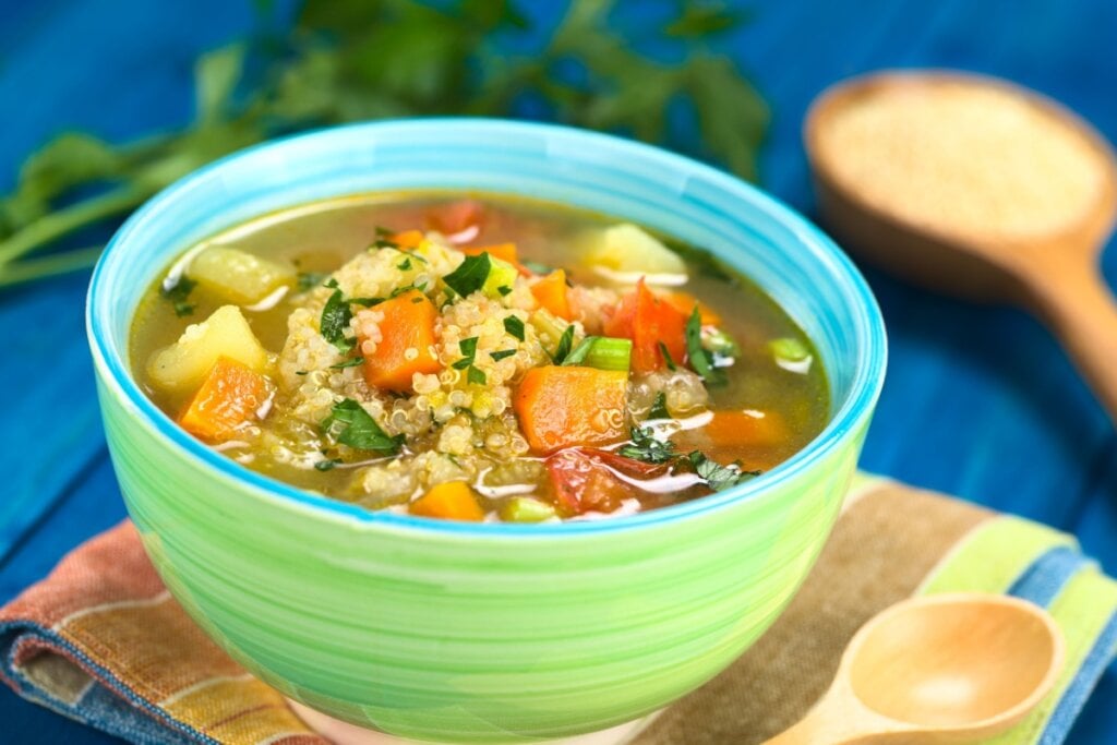7 sopas e cremes ricos em proteínas para incluir no cardápio 6 Sopa de quinoa com legumes servido em tigela de cerâmica verde em cima pano de prato listrado em cima de mesa de madeira azul