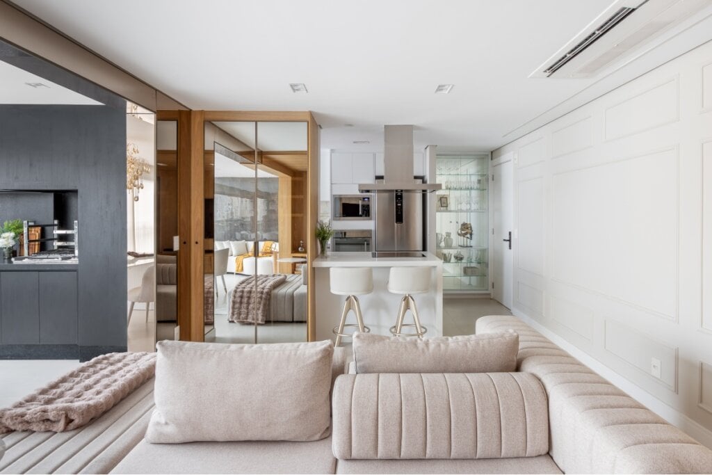 3 materiais que elevam a atmosfera dos ambientes 11 Sala de estar com um sofá bege na frente e, ao fundo, um móvel marrom com vidro e uma cozinha